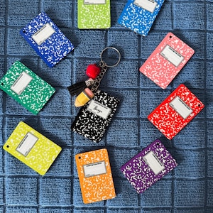 Puede incluir: Una colección de llaveros de libretas de composición de colores, incluyendo verde, azul, rosa, rojo, amarillo, naranja y morado. Un llavero presenta un charm de libreta negra con la palabra "Things" y una borla.