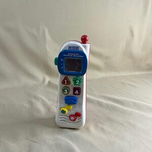 May include: A white and red VTech Tiny Touch Phone Plus toy with a blue screen, green button, and colorful buttons. The phone has a red antenna and features the numbers 1, 2, 3, and 4.