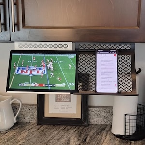 Peut inclure: Un support métallique blanc et noir pour sous-meuble, maintenant une tablette affichant un match de football NFL et un smartphone montrant une liste de texte. Le support présente un panneau arrière en treillis et une étagère plate.