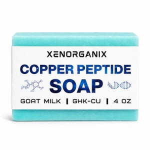 Puede incluir: Una pastilla de jabón rectangular de color azul claro envuelta en una etiqueta de papel blanco. La etiqueta dice XENORGANIX COPPER PEPTIDE SOAP con un gráfico de estructura química y hélice de ADN. El texto incluye GOAT MILK, GHK-CU y 4 OZ.