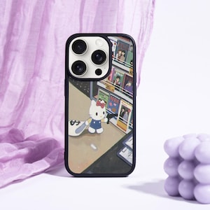 May include: Black phone case featuring a colorful illustration of Hello Kitty in a city scene. The case has a black border and a glossy finish. A lavender bubble candle and a purple fabric are in the background.