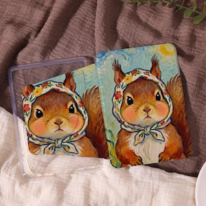 May include: Two decorative cards featuring a squirrel wearing a floral headscarf. The squirrel has a light brown coat, rosy cheeks, and a serene expression. One card is inside a clear plastic case. The background of the cards is a light blue sky with a yellow sun.