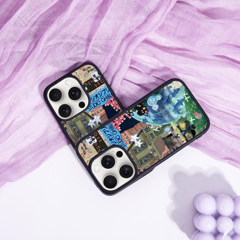 May include: Two black phone cases with a colorful collage design featuring Japanese text and illustrations. The cases have a black border and a camera cutout. The design includes images of buildings, characters, and other graphics.