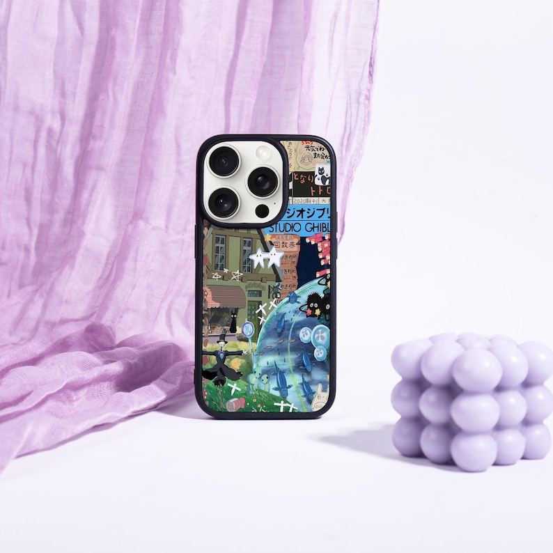 May include: A black phone case featuring a colorful collage of Studio Ghibli artwork. The design includes images of buildings, characters, and Japanese text. A lavender bubble candle is in the background.