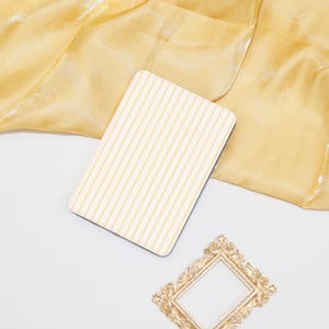 May include: A cream-coloured card with vertical stripes, a gold-coloured ornate picture frame, and a piece of gold-coloured fabric. The card is on a white surface, with the fabric and frame nearby.