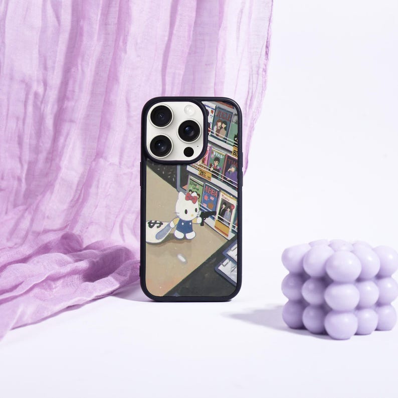May include: Black phone case featuring a colorful illustration of Hello Kitty in a city scene. The case has a black border and a glossy finish. A lavender bubble candle and a purple fabric are in the background.