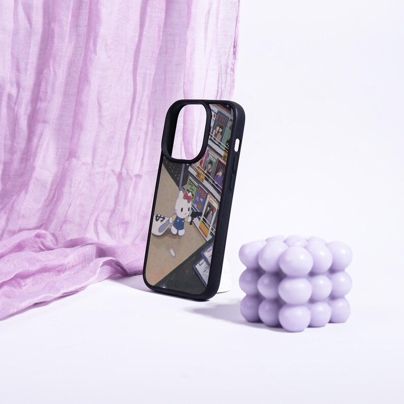 May include: Black phone case featuring a colorful illustration of Hello Kitty with a skateboard in front of a store. A lavender bubble candle and a draped fabric are also in the image.