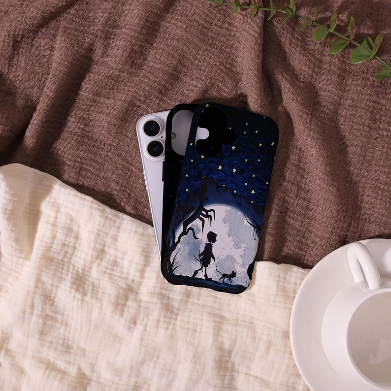 May include: A black phone case featuring a silhouette of a person, a cat, and a tree in front of a full moon. The case has a dark blue night sky with yellow stars. A silver phone is next to the case.