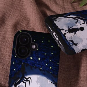May include: Two phone cases with a dark blue night sky design, featuring a full moon, stars, and a silhouette of a figure. One case shows the camera lenses. The other case has a silhouette of a figure in a tree.