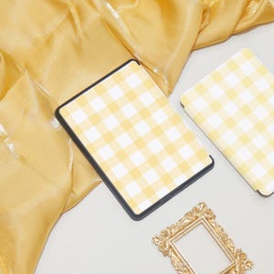 K&ouml;nnte beinhalten: Eine gelbe und wei&szlig;e karierte E-Reader-H&uuml;lle mit einem goldenen Bilderrahmen auf einem gelben Stoffhintergrund.