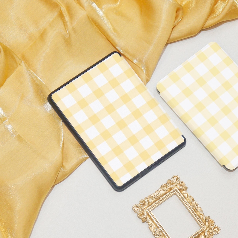 K&ouml;nnte beinhalten: Eine gelbe und wei&szlig;e karierte E-Reader-H&uuml;lle mit einem goldenen Bilderrahmen auf einem gelben Stoffhintergrund.
