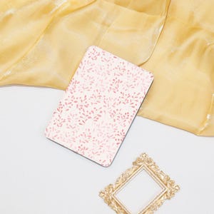 Puede incluir: Una cubierta de cuaderno blanca con un patrón floral rosa. La cubierta está sobre un fondo de tela dorada.