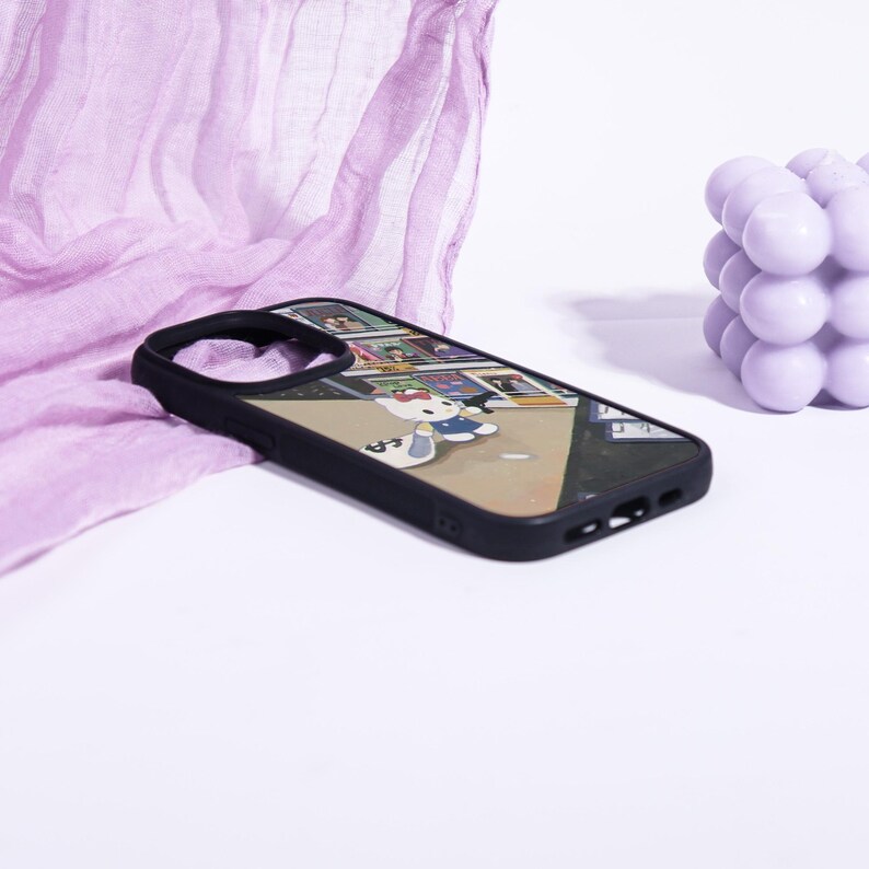 May include: Black phone case featuring a cartoon illustration of Hello Kitty in a blue dress. The case has a black border and a colorful background. The phone case is lying on a white surface with a purple cloth and a purple bubble candle in the background.