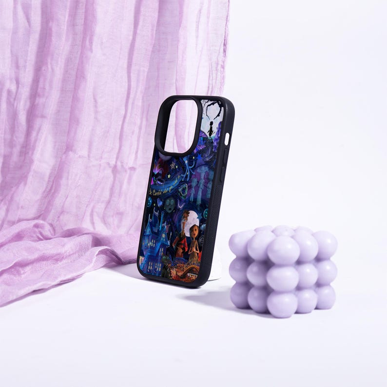 May include: Black phone case with a colorful illustration featuring a fantasy scene with characters and a castle. A lavender bubble candle sits next to the phone case. A lavender fabric is in the background.