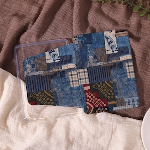 Può includere: Una custodia in plastica trasparente con un inserto in tessuto patchwork blu. Il tessuto presenta vari motivi, tra cui quadrati, strisce e motivi floreali nei toni del blu, marrone e rosso. La custodia è aperta.