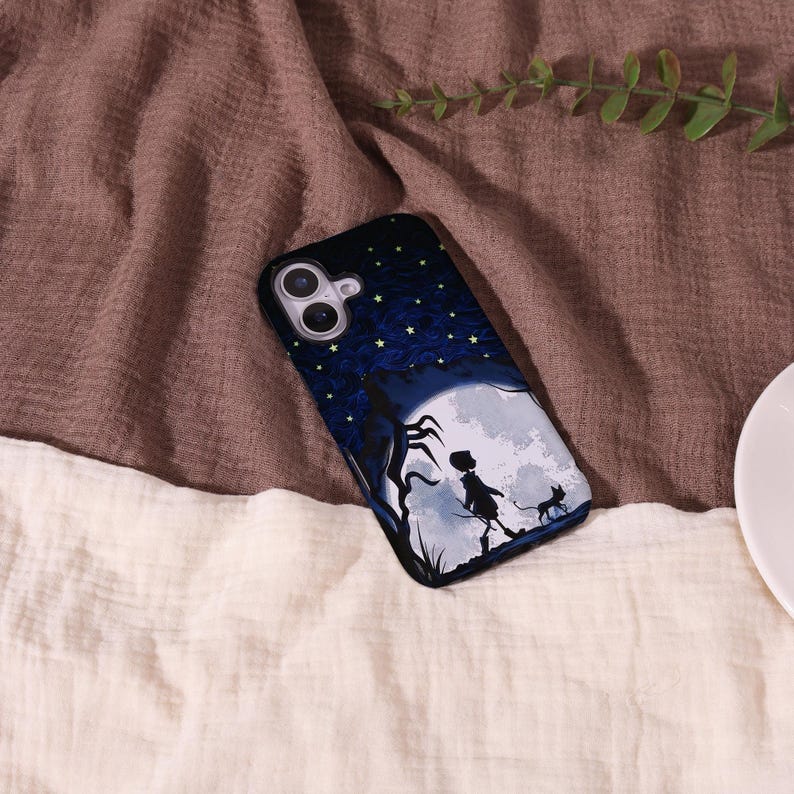 May include: A black phone case featuring a whimsical illustration of a silhouetted figure, a cat, and a tree against a full moon and starry night sky. The top of the case has a dark blue background with yellow stars.