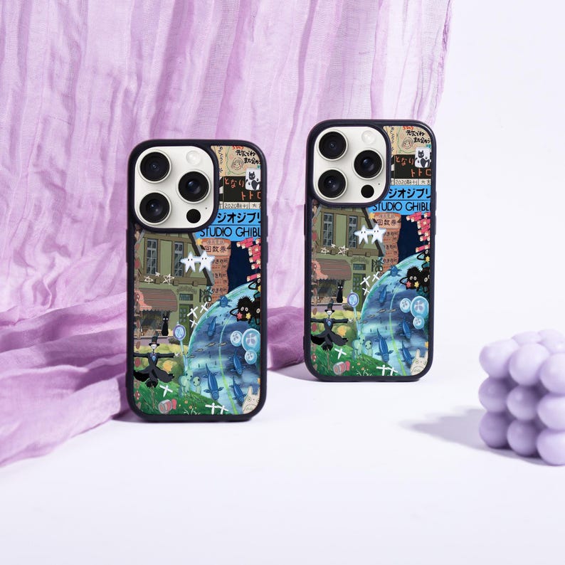 May include: Two black phone cases with a colorful Studio Ghibli design. The cases feature a collage of illustrations, including buildings, water, and Japanese text. The cases have a black border and are set against a purple fabric background.