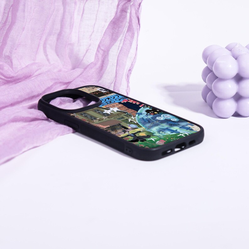 May include: Black phone case with a colorful graphic design. The case features a collage of images, including buildings, text, and a blue and green illustration. The case is set against a white background with a purple fabric and a lavender bubble-shaped object.