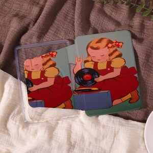 Può includere: Una custodia per telefono trasparente e una scheda abbinata con un'illustrazione vintage di una ragazza in un vestito rosso con un fiocco, che tiene in mano un disco in vinile. Lo sfondo è azzurro con un'estetica retrò.