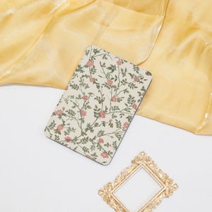 Op de afbeelding: Een rechthoekig notitieboek met een bloemenontwerp, met roze rozen en groene bladeren op een crèmekleurige achtergrond. Het notitieboek ligt op een gele stof, met een gouden decoratief frame op de voorgrond.