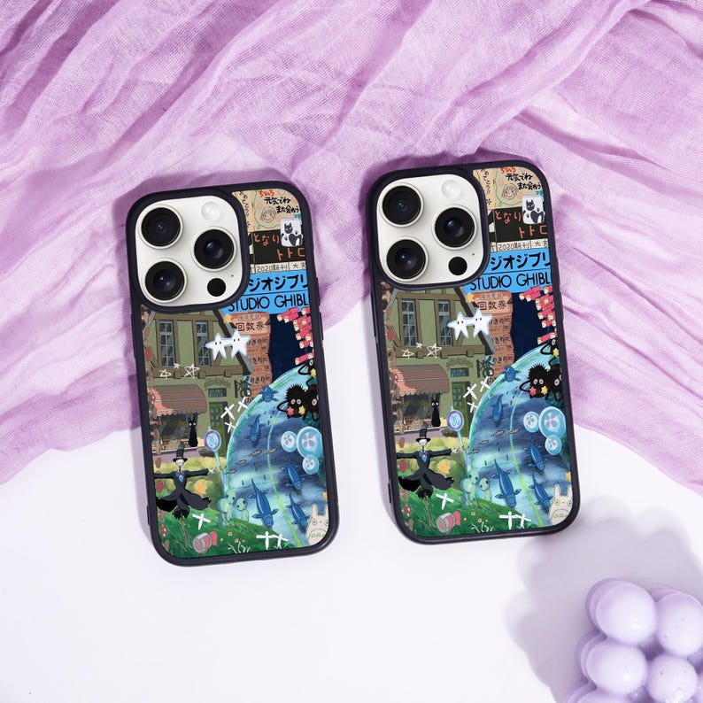 May include: Two black phone cases with a colorful illustration featuring a whimsical landscape, characters, and Japanese text. The cases have a black border and a camera cutout. The cases are on a purple fabric surface.