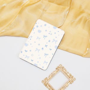 Op de afbeelding: Een witte notitieboekomslag met een blauw en wit patroon van strikken, harten en sterren. De notitieboekomslag ligt op een gele stoffen achtergrond.