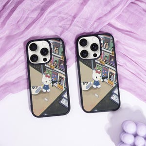 May include: Two black phone cases with a cartoon illustration of Hello Kitty in a city scene. The cases have a black border and a design featuring Hello Kitty holding a bag of money. The background shows a street with shops.