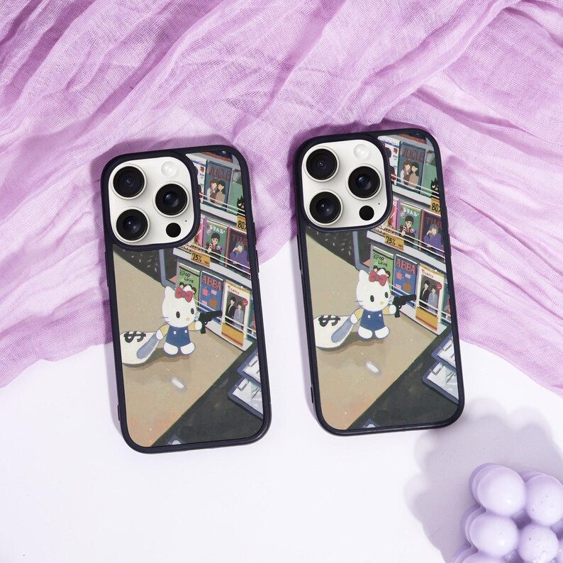 May include: Two black phone cases with a cartoon illustration of Hello Kitty in a city scene. The cases have a black border and a design featuring Hello Kitty holding a bag of money. The background shows a street with shops.