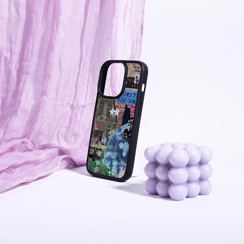 May include: Black phone case with a colorful collage design featuring Japanese text and illustrations. A light purple bubble candle sits next to the phone case. The background includes a light purple fabric.