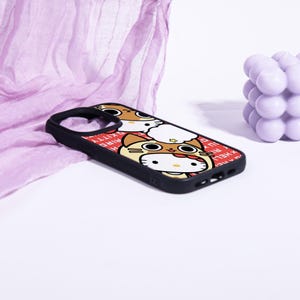 May include: Black phone case featuring a cartoon design with a cat wearing a costume. The case has a red border with white text. The phone case is on a white surface with a purple cloth and a purple decorative object in the background.