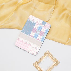 May include: A rectangular, patterned notebook with a patchwork design in pastel pink, blue, and white. The notebook features floral lace trim and a gold-colored decorative frame.