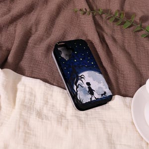 May include: A black phone case featuring a whimsical illustration of a person and a cat walking towards a large moon under a starry night sky. The case has a silver border and a dark blue background.