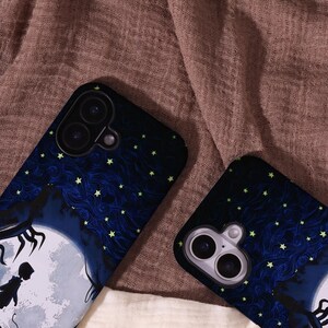 May include: Two phone cases with a dark blue and black design featuring a full moon, silhouette figures, and a starry night sky. The cases have a glossy finish and are set against a brown fabric background.