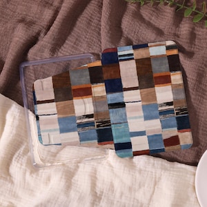 Può includere: Una custodia rettangolare con un inserto fantasia colorato. L'inserto presenta un design patchwork con quadrati blu, marroni e beige. La custodia trasparente è aperta, rivelando l'inserto. Lo sfondo è un tessuto marrone strutturato.