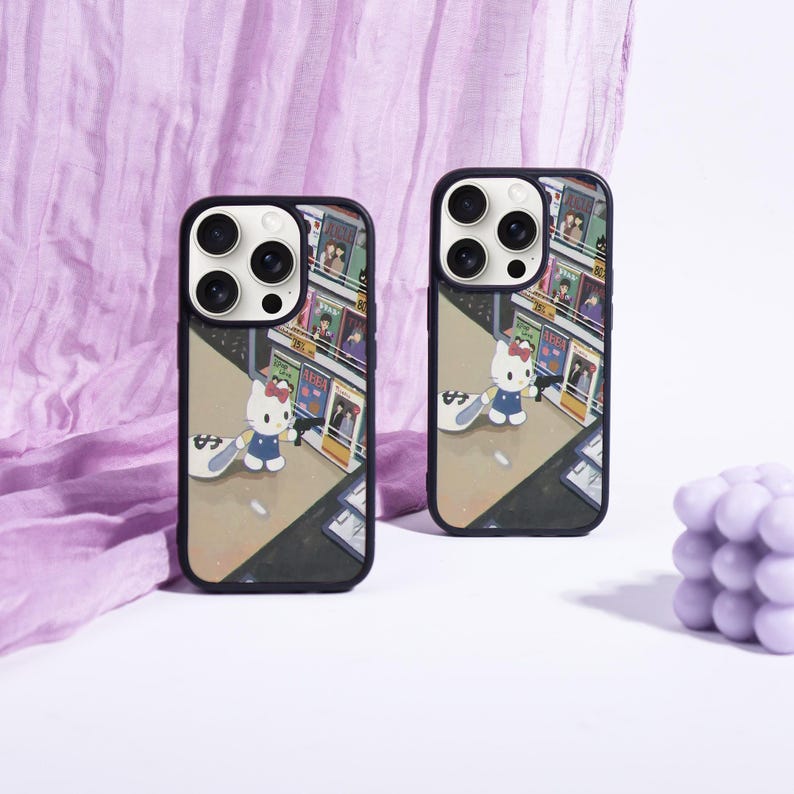 May include: Two black phone cases with a colorful illustration of a street scene featuring Hello Kitty. The cases have a black border and a camera cutout. The background includes a purple fabric and a lavender-colored decorative object.