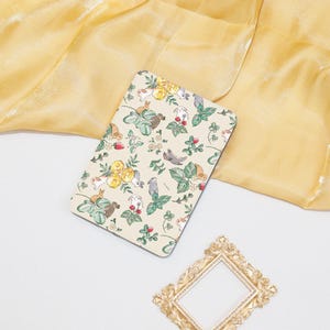 Può includere: Un taccuino rettangolare color crema con un motivo stravagante di conigli, fiori e fragole. Il taccuino è appoggiato su uno sfondo di tessuto giallo, con una cornice decorativa dorata nell'angolo.