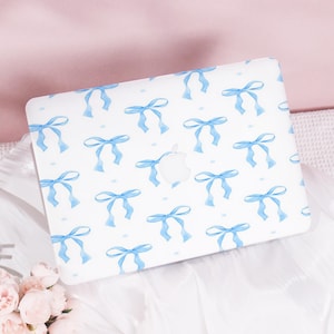 May include: White laptop case with a repeating pattern of light blue watercolour bows. The Apple logo is visible. The case is on a white surface with pink fabric in the background. The bows are evenly spaced across the surface.