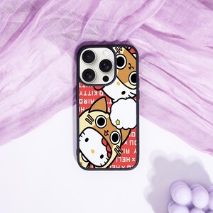 May include: Black phone case featuring a cartoon illustration of Hello Kitty in a shark costume. The case has a red background with white text. The phone case is on a purple surface.