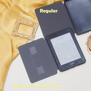 K&ouml;nnte beinhalten: Ein grauer Kindle E-Reader mit schwarzem Cover und einem Handriemen. Der Bildschirm zeigt den Text "La Chapitre VII" und das Wort "kindle".