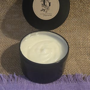 May include: A black jar with a white cream inside. The jar has a black lid with a brown label that says "God Gifted Resources" with a floral design and the letter "G".