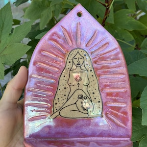 May include: A pointed arch-shaped ceramic wall plaque with a glossy iridescent pink glaze. The center features a black line drawing of a seated figure with long hair, a sun symbol on the chest, and a flower held in the lap, surrounded by rays.