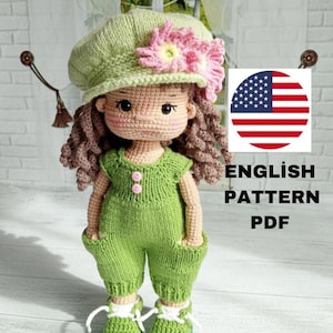 Può includere: Bambola all'uncinetto fatta a mano con una tuta verde, scarpe abbinate e un cappello verde chiaro ornato da un fiore rosa. La bambola ha i capelli ricci castani e un'espressione dolce. L'immagine include il testo "ENGLISH PATTERN PDF" e una bandiera americana.