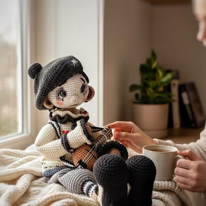 May include: A handmade crochet doll dressed as a musician, wearing a black hat with a white star, a white and black outfit, and black shoes. The doll is holding a small brown instrument. The doll is sitting on a cream-colored blanket.