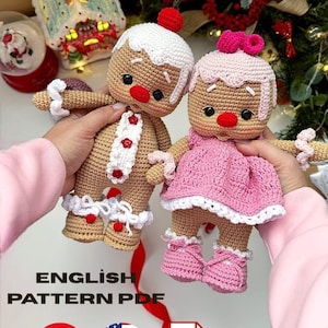 May include: Two crocheted gingerbread dolls with tan yarn bodies, red noses, and black eyes. One doll wears a white hat with a cherry and a white tie with red buttons. The other wears a pink dress, pink shoes, and a pink bow. Text reads ENGLISH PATTERN PDF.