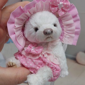 Op de afbeelding: Een handgemaakte witte teddybeer met een roze muts met een strik en een roze jurk met stippen. De beer heeft een lichtbruine neus en kleine zwarte ogen. De outfit bevat een bijpassende strik op de jurk. De beer is een pluche speelgoed.