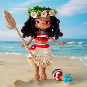 Op de afbeelding: Een gehaakte pop met bruin haar, die een bloemenkrans draagt, een rood-wit topje en een witte rok. De pop houdt een houten peddel vast en staat op een zandstrand.
