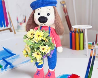 Puede incluir: Una muñeca de perro de ganchillo hecha a mano con boina azul, peto rojo y pantalón azul. El perro sostiene un ramo de flores amarillas y lleva zapatos rosas. La muñeca tiene orejas marrones y nariz negra.