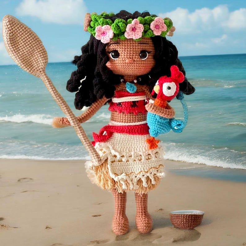 Op de afbeelding: Een gehaakte pop van Moana uit de Disney-film Vaiana, die een traditionele Polynesische outfit draagt met een bloemenkrans, en een blauw-rode gehaakte papegaai vasthoudt. De pop staat op een zandstrand met een kleine kokosnootschelp ernaast.