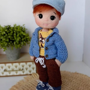 May include: A handmade crochet doll of a young boy. The doll has red hair, a blue cap, and a blue jacket over a yellow shirt with a graphic. He wears brown pants and blue shoes. The doll is standing on a woven surface.