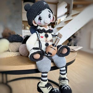 May include: A handcrafted crochet doll resembling a clown, with a black hat and a white and black outfit. The doll has grey trousers and holds a miniature ukulele. The doll's face features rosy cheeks and a playful expression.